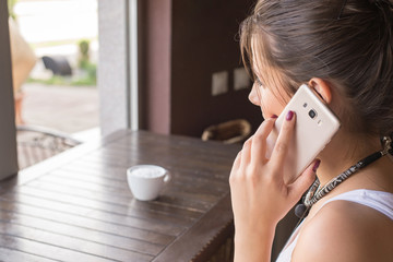 Girl talk to a cell phone in a coffee shop. View behind girl shoulder. Front of is table with a cup...