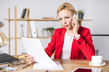 portrait of focused businesswoman with document talking on smartphone at workplace in office