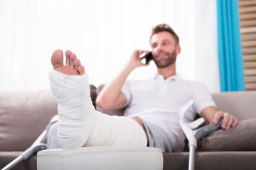 Happy Young Man With Broken Leg Talking On Smartphone