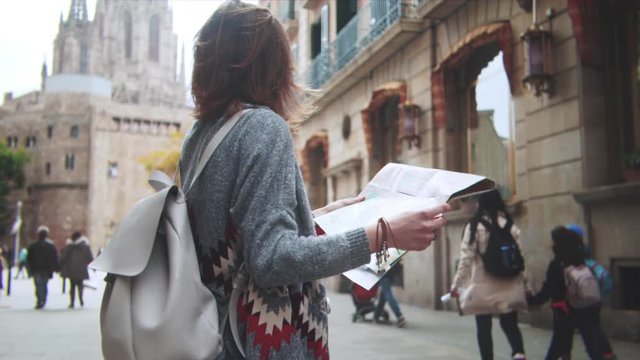 Back view of young hipster girl walking through European street and exploring Barcelona city with map, travel vacation adventure concept, slow motion