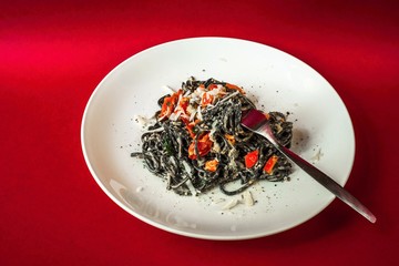 Black spaghetti with tomato and cheese on white plate on red background.