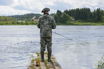 A fisherman with a fishing rod by the river. A slender man is on outdoor activity. A fisher is catching a fish on a wooden raft.