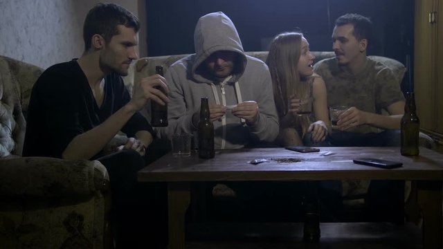 Four young alcoholics sitting on sofa at night in domestic room at home, abusing alcohol while hooded shirt male rolling marijuana joint. Alcohol abuse, social issues and drug addiction concept
