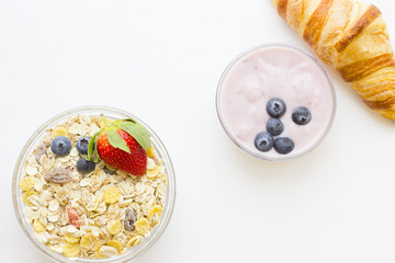 Healthy Breakfast Concept: a bowl of oat muesli with candied fruit and fresh strawberry and blueberries, a french croissant and youghurt on white, top view, copy space