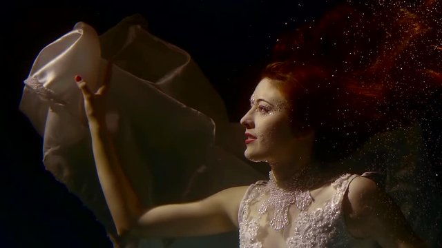 Charming young woman with a red hair is posing with her floating white dress underwater.