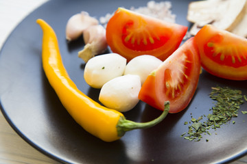Preparing Food Concept. Ingredients for a meal: tomato, chili pepper,garlic, mozarella, herbs and spices on a black plate, white wooden background