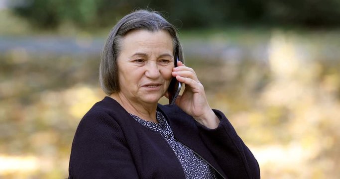 Positive senior woman talking on smartphone outdoors in park.