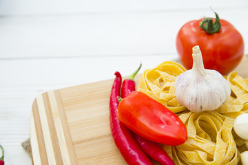 Healthy Food, Kitchen Concept. A set of cooking ingredients on a wooden cutting board: fetuccini nests, tomato, chilli peppers, garlic and mozarella cheese, white wooden background, copy space