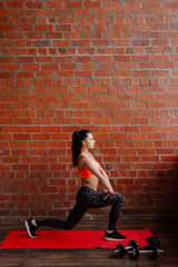 Fototapeta na wymiar Young, fit and sporty woman in leggings and top in front of brick white wall. The woman trains at home, does warm-up and an extension. Fitness, sport lifestyle concept.