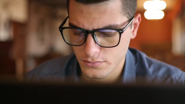 Close-up of reflections of laptop monitor screen in man's glasses. Young caucasian freelancer businessman working and browsing internet in office. Hacker at work stealing accounts database.