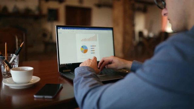 Back view of freleancer businessman in glasses diligently working on laptop in cafe. Man typing on keyboard of computer with diagrams and trends document on the screen. Business concept