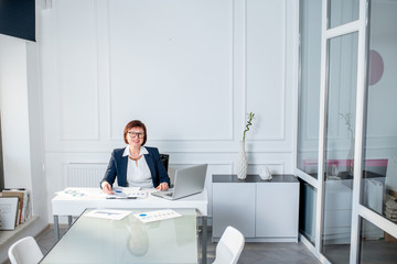 Elegant senior businesswoman dressed in the suit working with laptop and documents at the white office. Wide angle view with copy space