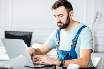 Handsome repairman or foreman in uniform working with laptop and architectural drawings at the...
