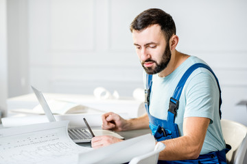 Handsome repairman or foreman in uniform working with laptop and architectural drawings at the office