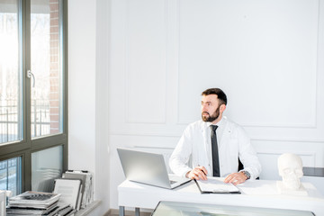 Fototapeta na wymiar Handsome male doctor in the medical gown working with laptop sitting in the beautiful white office interior