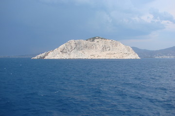 Panorama of a small Greek island surrounded by waves of the Aegean Sea and against the background of a cloudy blue sky at sunset.