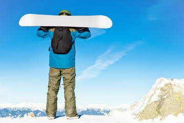 Winter sports concept - male snowboarder holding his snowboard on his shoulders at the top of a mountain and enjoying the scenery - outdoors shot