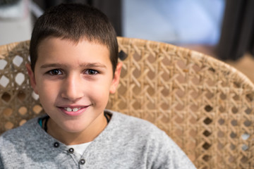 Beautiful happy young boy sitting in the knitted chair, looking at camera, smiling 
