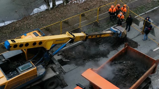 The excavator digs the asphalt road, a group of workers looks at the process, 4K.