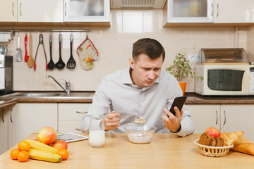 Handsome young business man in plaid shirt having breakfast, sitting at table, talking on mobile phone, eating cereals with milk on light kitchen. Healthy lifestyle. Cooking at home. Prepare food.