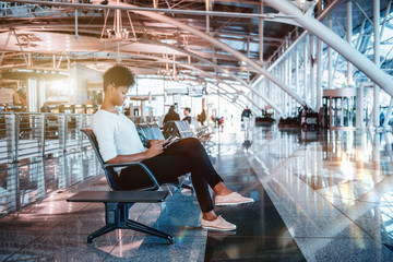 Pensive young Brazilian tourist female is sitting inside of modern waiting hall of airport terminal and waiting for her flight; cute black girl is waiting indoors of contemporary railway station depot