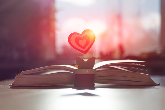 symbol of love red heart in the form of a flower stands near an open old book on a background of a blurred window in the yellow rays of the sun