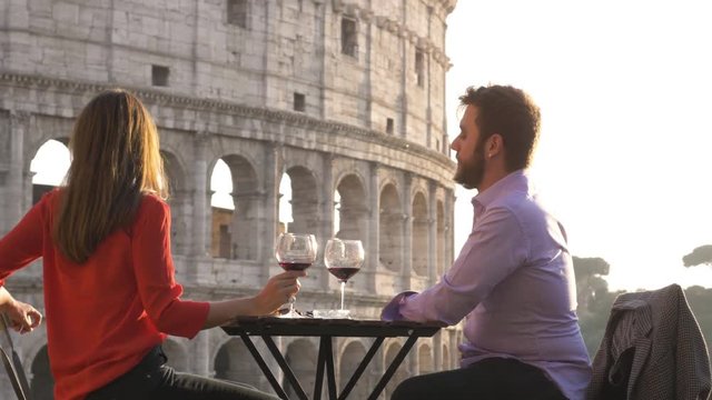 Romantic elegant young couple marriage proposal with ring sitting at restaurant in front of colosseum in rome at sunset with cup of red wine emotional