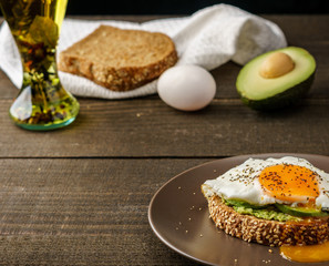 sandwich with avocado and a fried egg on a brown plate on a rustic wood and table bread on white kitchen towel.