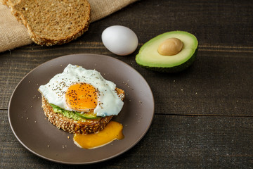 sandwich with avocado and a fried egg on a brown plate on a rustic wood and table bread on sackcloth.