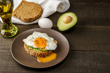 sandwich with avocado and a fried egg on a brown plate on a rustic wood and table bread on white kitchen towel.