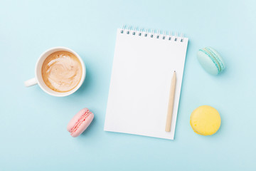 Clean notebook, cup of coffee and colorful macaron on blue desk top view. Cozy morning breakfast. Fashion flat lay. Sweet macaroons.
