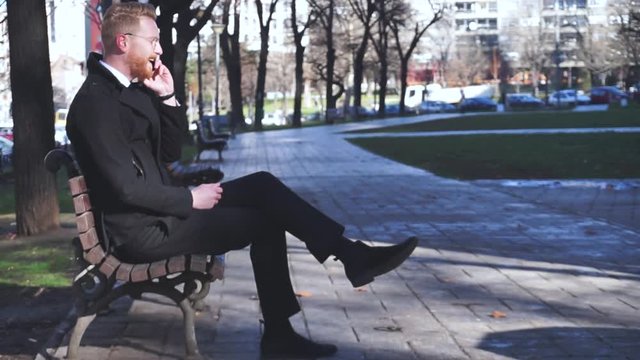 Young ginger businessman sitting alone in park and making a phone call
