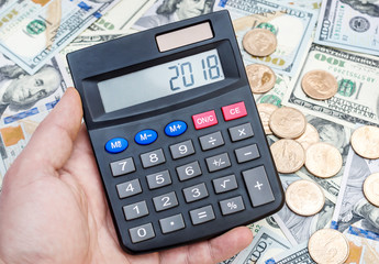 Hand holding calculator with numbers 2018 on display over background of dollars. Business concept.
