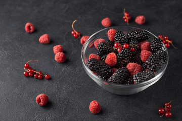 Glass plate with ripe red blackberries, raspberries and currant on a black surface.