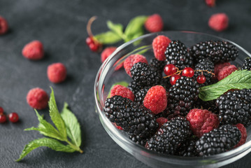 Glass plate with ripe red blackberries, raspberries, currant and green mint leaves on a black surface.