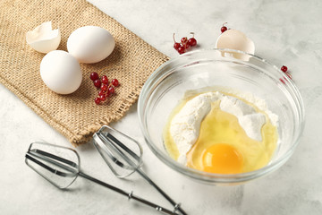 View at a plate of flour with brocken egg. Eggs and ripe red berries with egg shell on a bagging on a white background. Mixer fits a the foreground.
