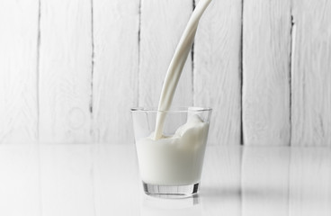 Milk is poured into the glass on a white surface infront of a white wooden background.