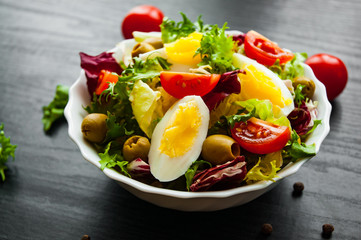 various fresh mix salad leaves with tomato, eggs and olive in bowl on dark wooden background
