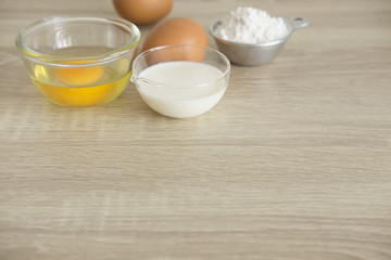 BOARD WITH BAKERY INGREDIENTS WITH WRITING SPACE
Important bakery ingredients, white flour in a measuring cup,  milk and eggs in transparent cups are on a wood table.