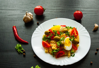 fusilli pasta salad with tomato, eggs and olive in plate on dark wooden background