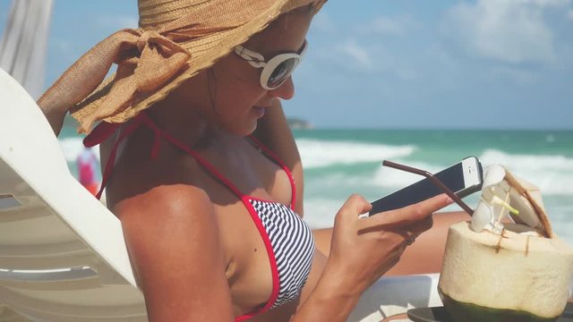 Young pretty brunette woman wearing sunglasses and hat lies on a lounger by the sea uses mobile phone. 3840x2160