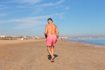 Rear view of unrecognizable shirtless senior man walking on empty sandy beach of Valencia, Spain in winter.