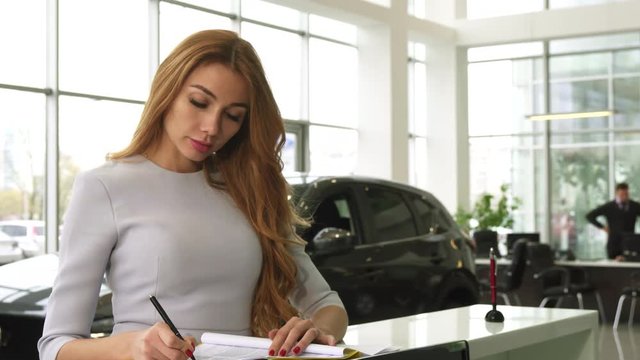 Young gorgeous woman buying a new auto at the dealership signing papers smiling to the camera happily consumerism retail rental buyer shopper shopaholic luxury ownership driving insurance.