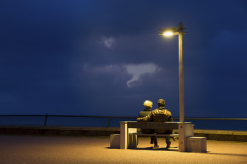 A woman and a man look at the sea sitting on a bench at dusk