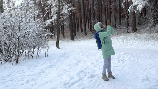 Beautiful young woman in winter clothing, mittens and wool hat photographing snowy forest by a knitted camera