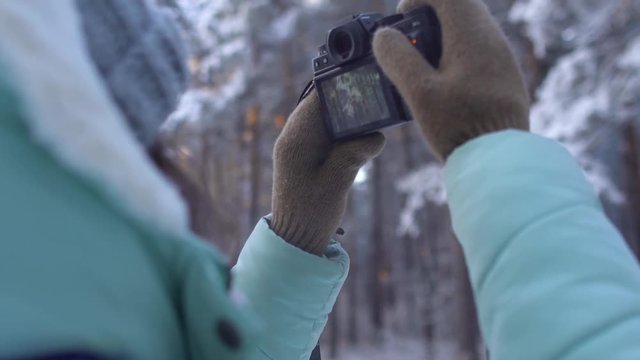 Beautiful young woman in winter clothing, mittens and wool hat photographing snowy forest by a knitted camera