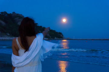 back view beautiful asian woman point finger to full moon over the seascape and mountain with reflection on the water surface at night.