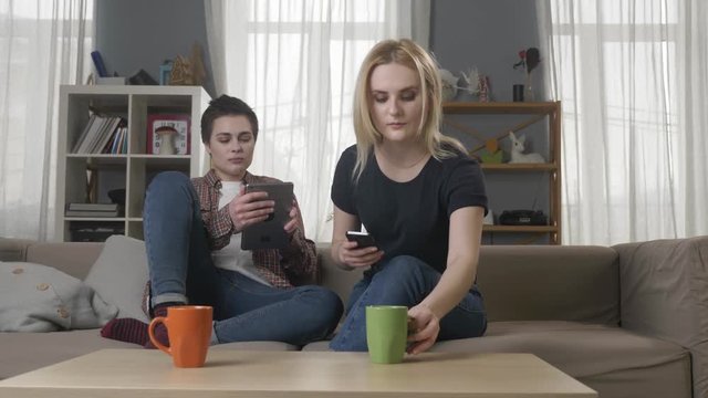 Two young girls are sitting on the couch and using a smartphone and a tablet, lesbians, a girl with short hair shows a funny photo on a tablet to her friend 60 fps