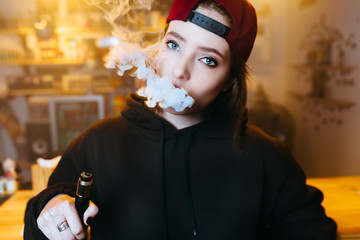 Young pretty woman in red cap smoke an electronic cigarette at the vape shop. Hip-hop style....