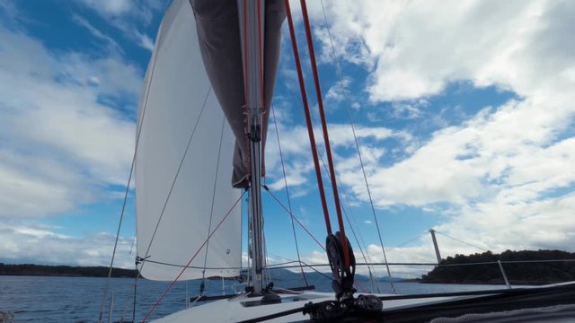 Movement of sailing boat on sea. Background large bridge across lake, river, horizon, high mountains and clear blue sky. View of ropes, mast, sails in bow of boat. Sails flutter from wind. POV shot